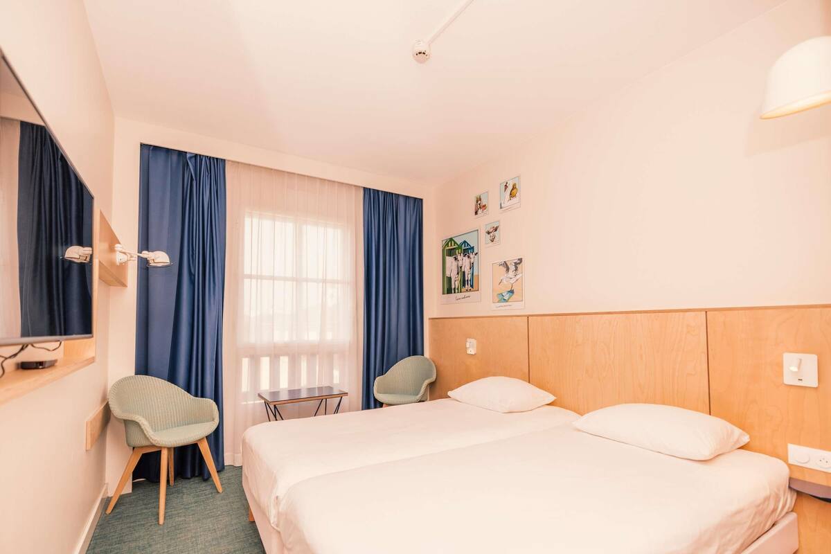 A hotel room with a bed, two chairs, a small table, a mirror, and a window with curtains.