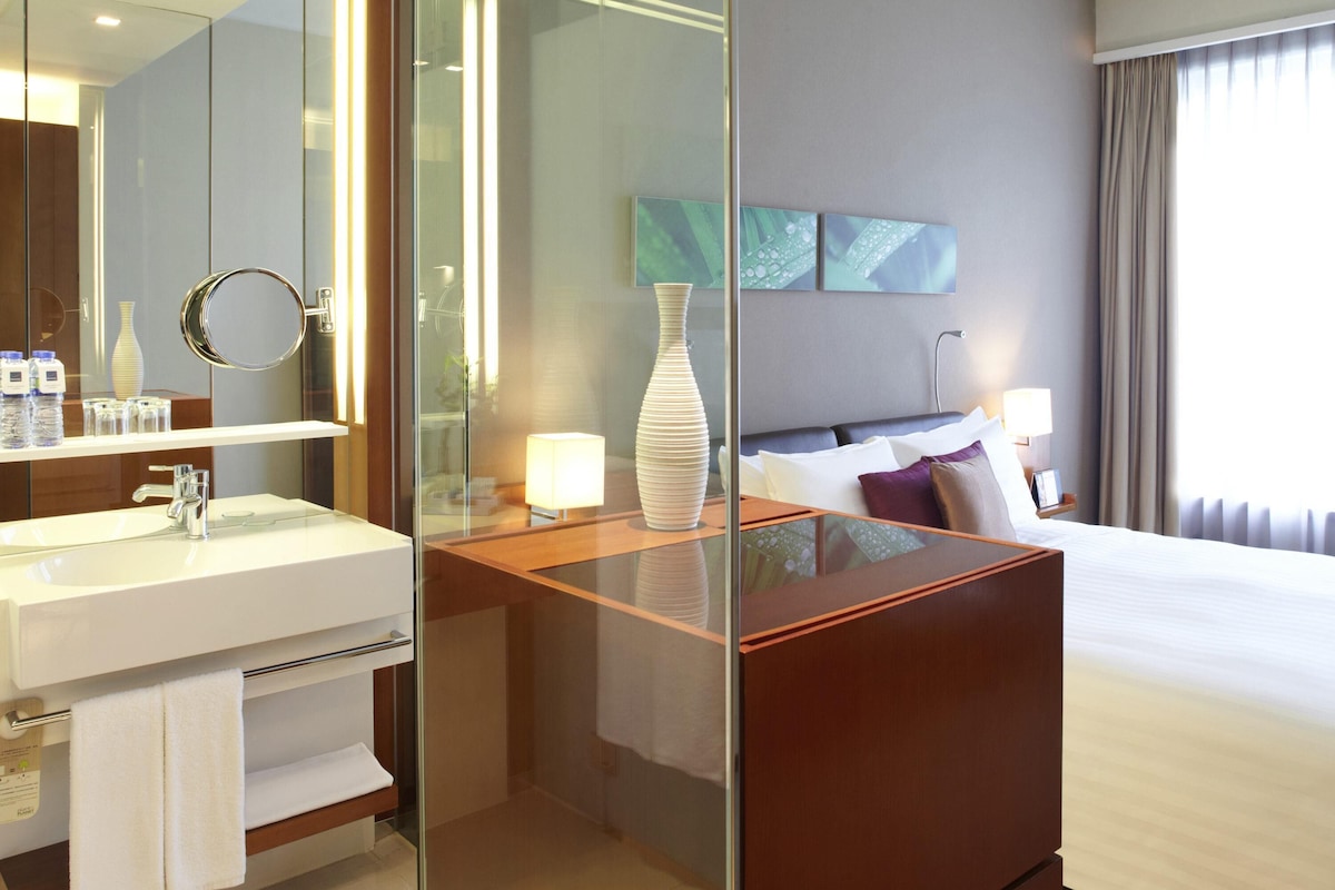 Une chambre d’hôtel moderne dotée d’un coin toilette vitré, d’un grand lit, d’une table de chevet avec une lampe et offrant une vue sur l’extérieur.