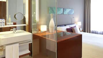 A modern hotel room with a glass-enclosed vanity, a large bed, a bedside table with a lamp, and a view of the outdoors.
