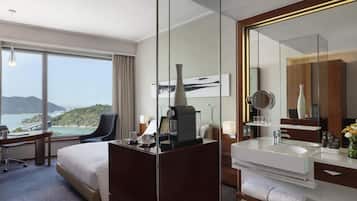 A hotel room with a bed, desk, chair, and a large window offering a view of the sea and hills.