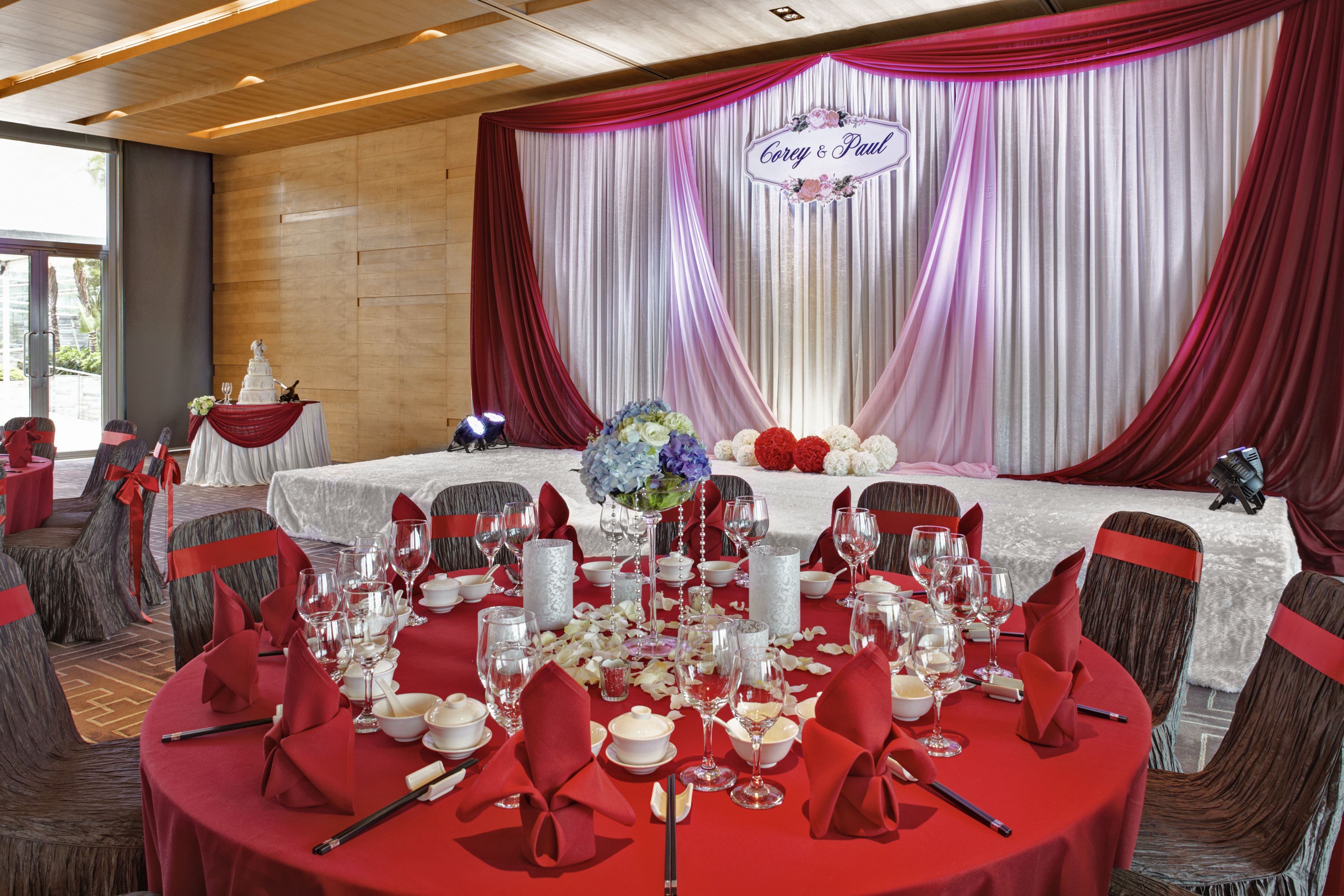 Un salón de banquetes con mesas redondas dispuestas para un evento formal, que incluye un escenario con fondo decorativo y un cartel que dice "Corey & Paul".