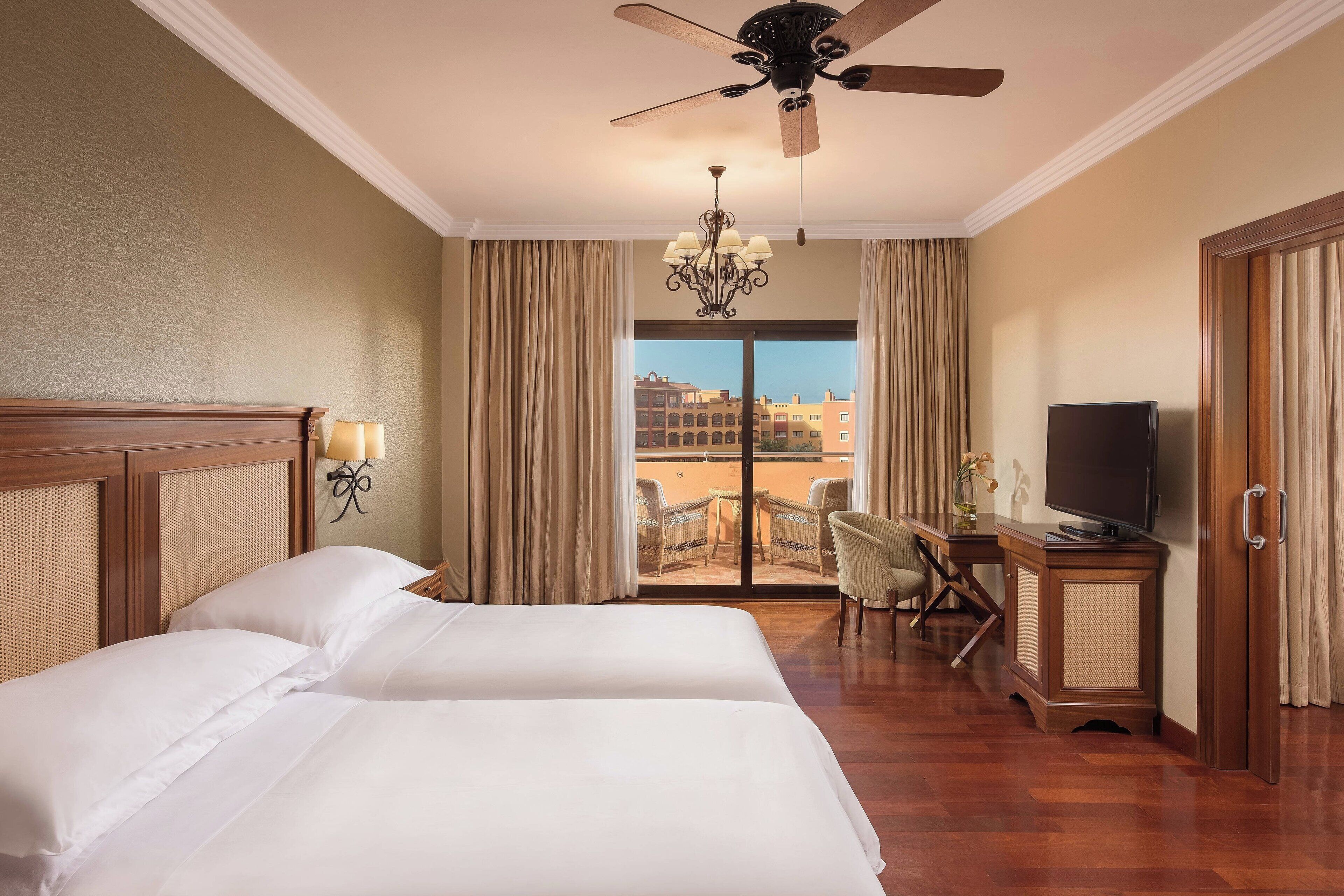 A hotel room with two beds, a ceiling fan, a chandelier, a television, a desk with a chair, and a view of buildings through the window.