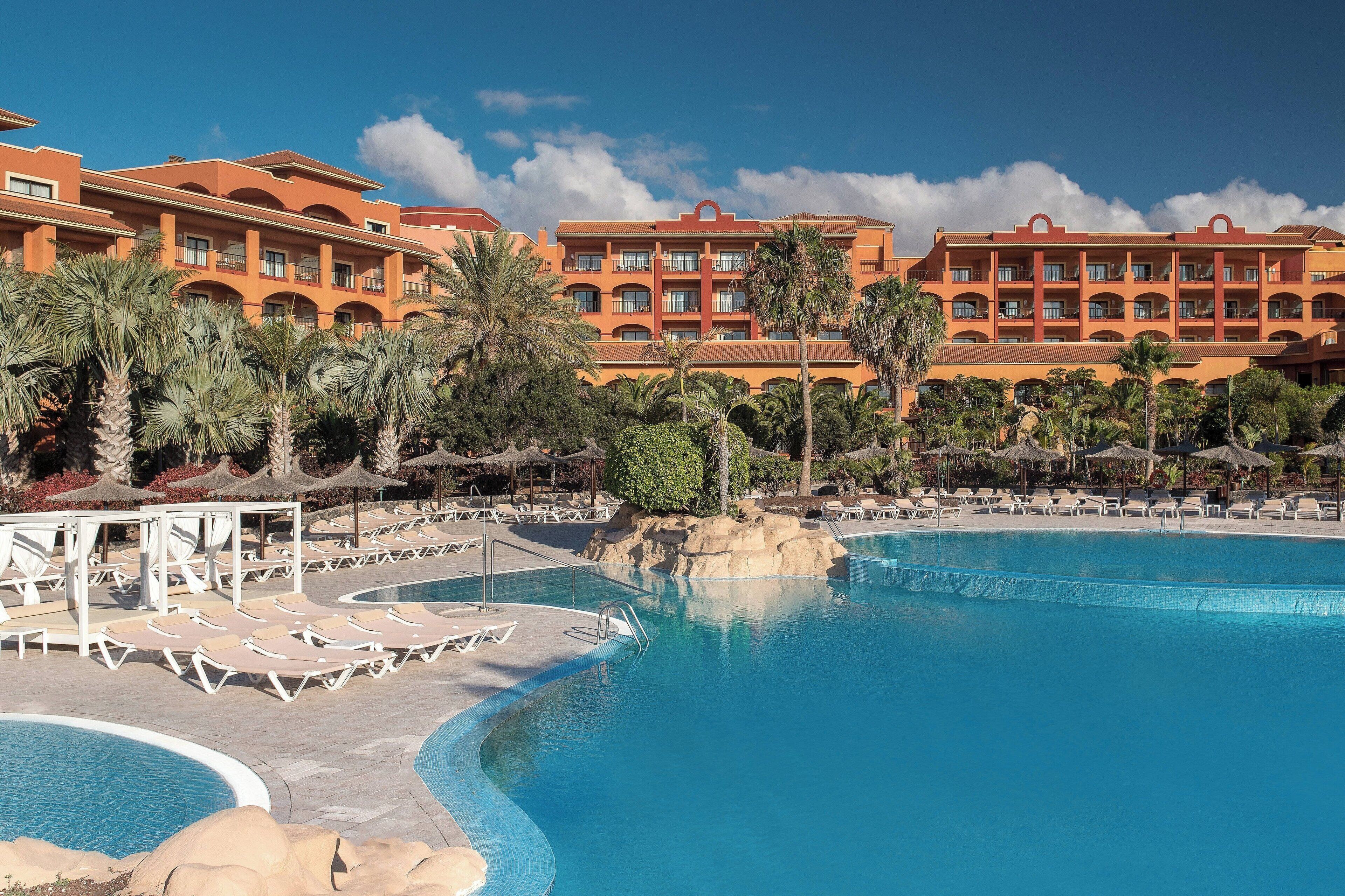 Un hotel con una piscina grande, palmeras y un cielo azul despejado.