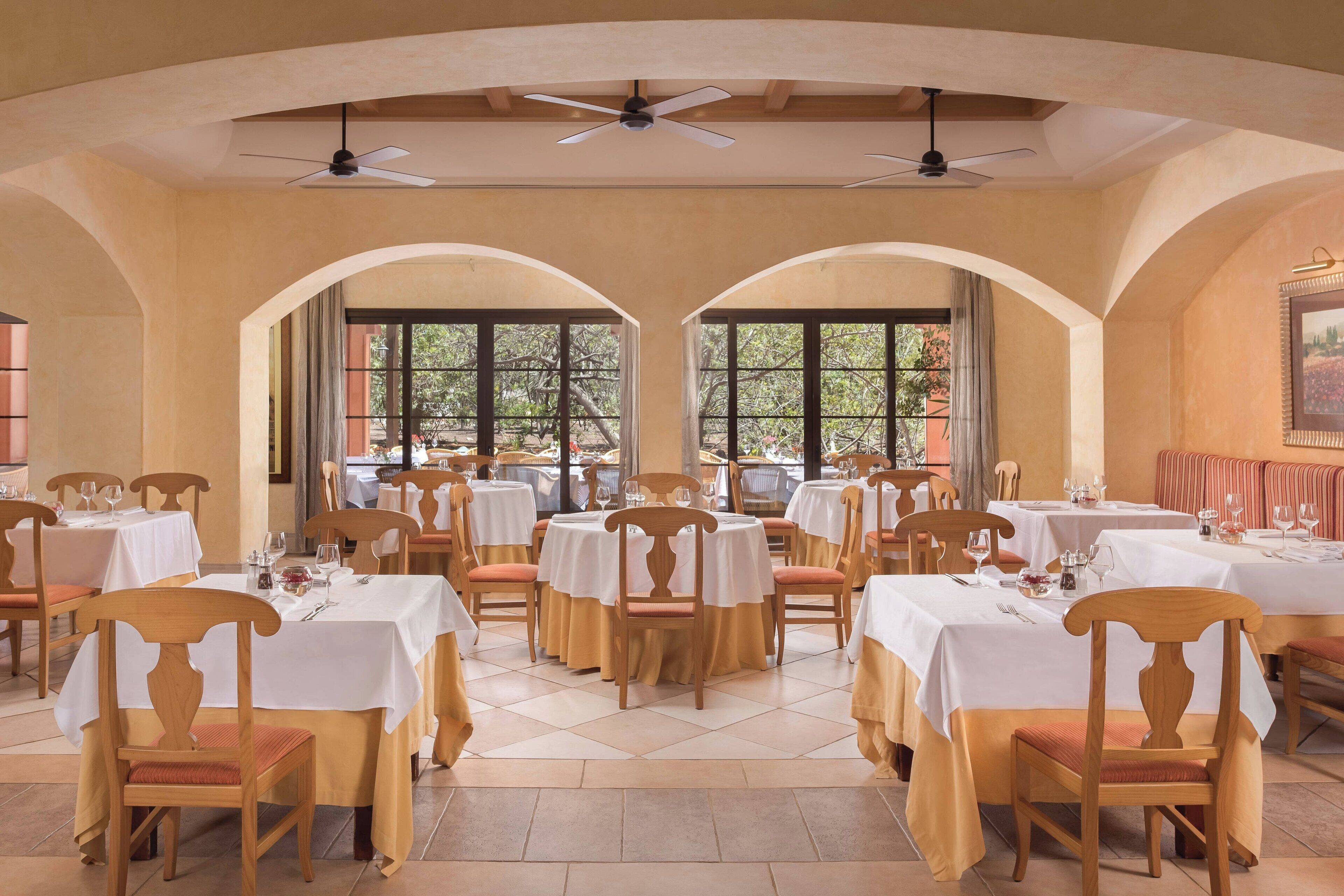 Un comedor con varias mesas y sillas, ventanas amplias con vistas a árboles y ventiladores de techo.