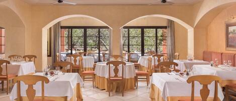 Un comedor con varias mesas y sillas, ventanas amplias con vistas a árboles y ventiladores de techo.