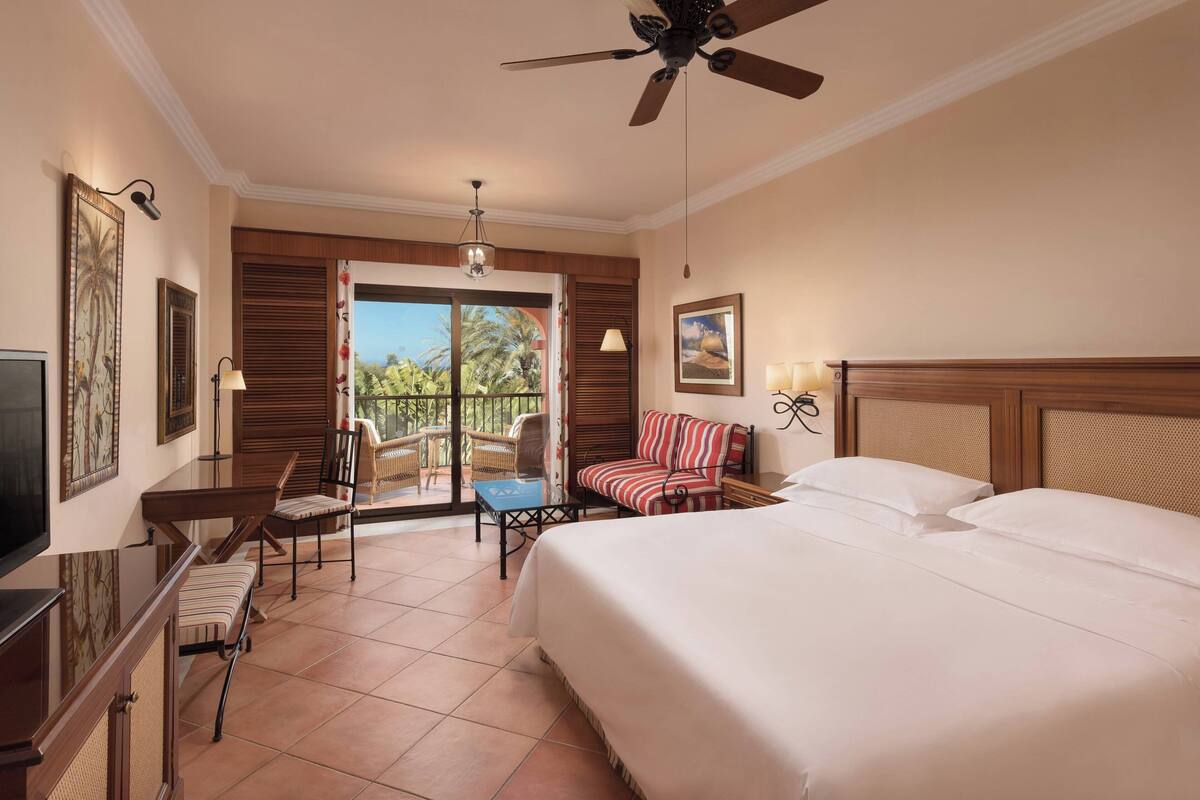 Habitación de hotel con cama, escritorio, silla, mesita, ventilador de techo y balcón con vistas a palmeras.