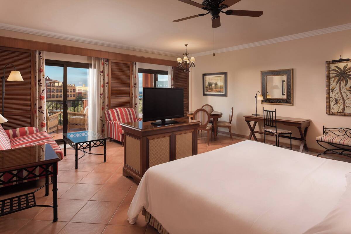 Habitación de hotel con una cama grande, televisión, escritorio y balcón con mobiliario de mimbre.