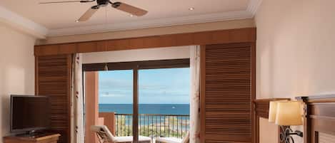 Habitación de hotel con cama, televisión, ventilador de techo, balcón con vista al mar y una lámpara.
