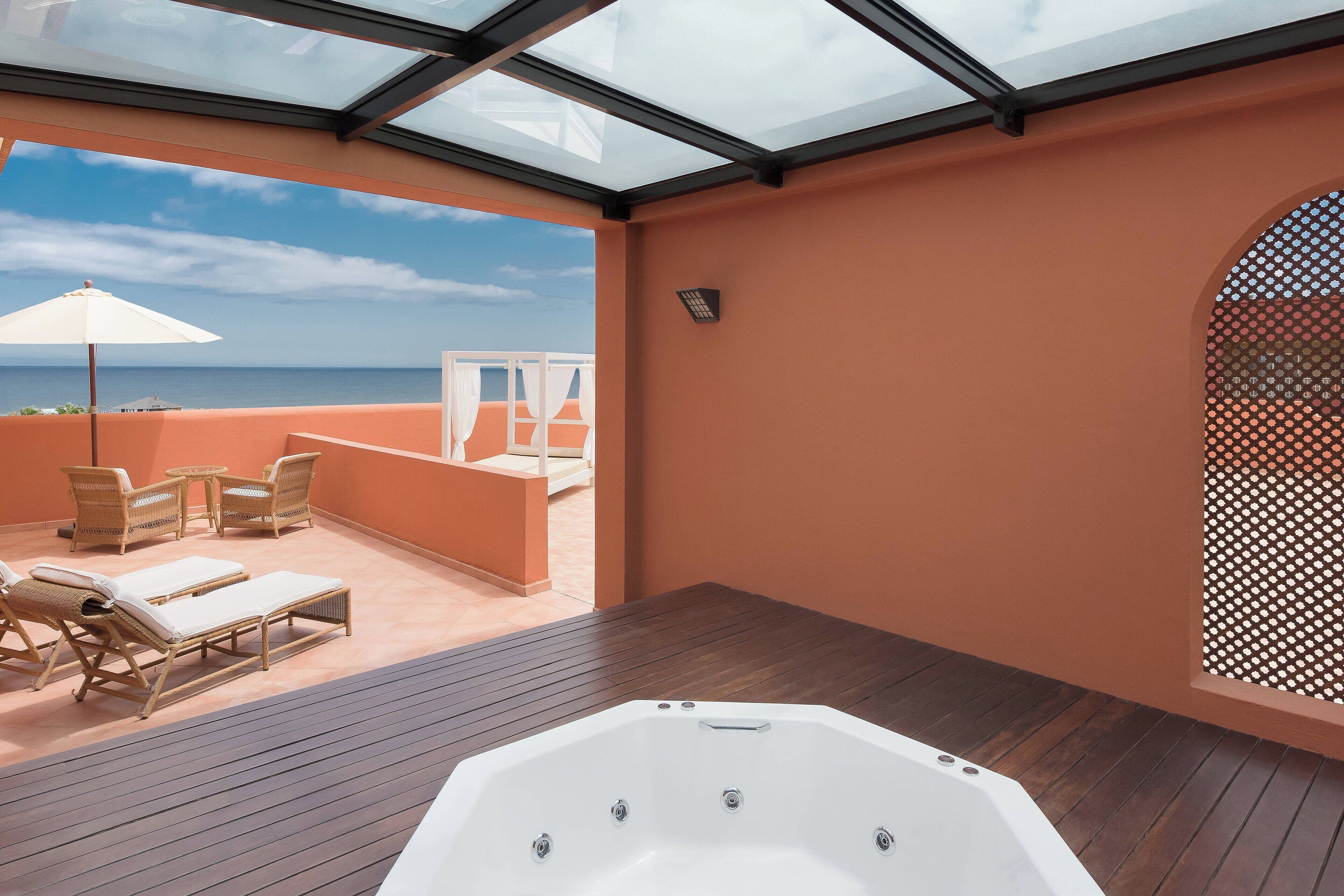 Een zonnige kamer met een houten vloer, een groot wit bad, rieten lounge stoelen en uitzicht op zee.