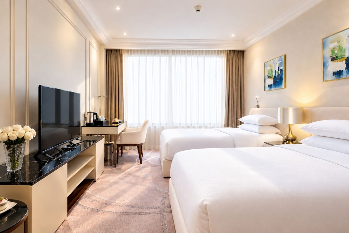 A hotel room with a flat-screen TV, a desk with a chair, two beds, a bedside table with a lamp, and a painting on the wall.