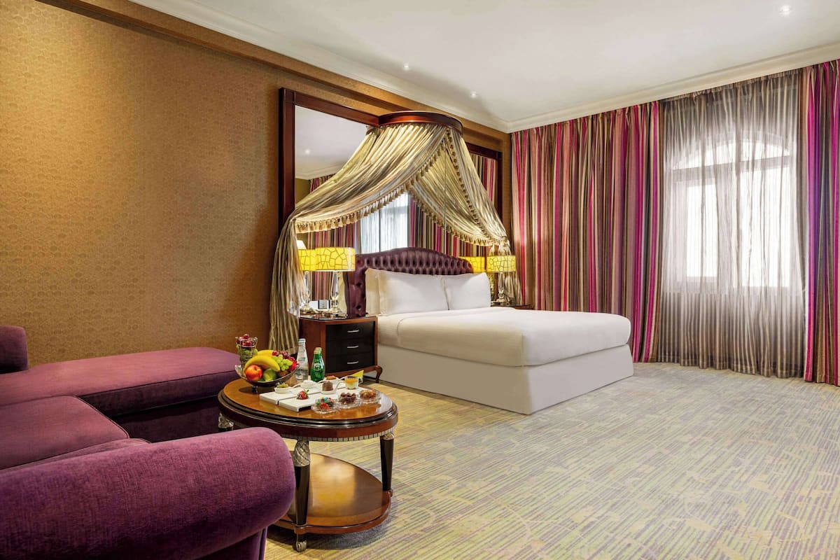 Una habitación de hotel con cama a dosel, una mesa redonda con fruta y bebidas, un sofá morado y cortinas a rayas.