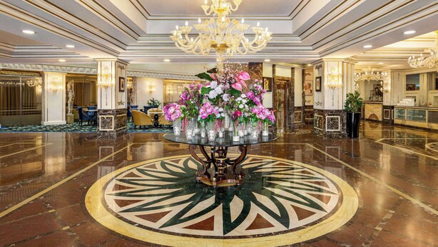 A grand lobby with a central round table adorned with flowers, a chandelier, and a polished marble floor.