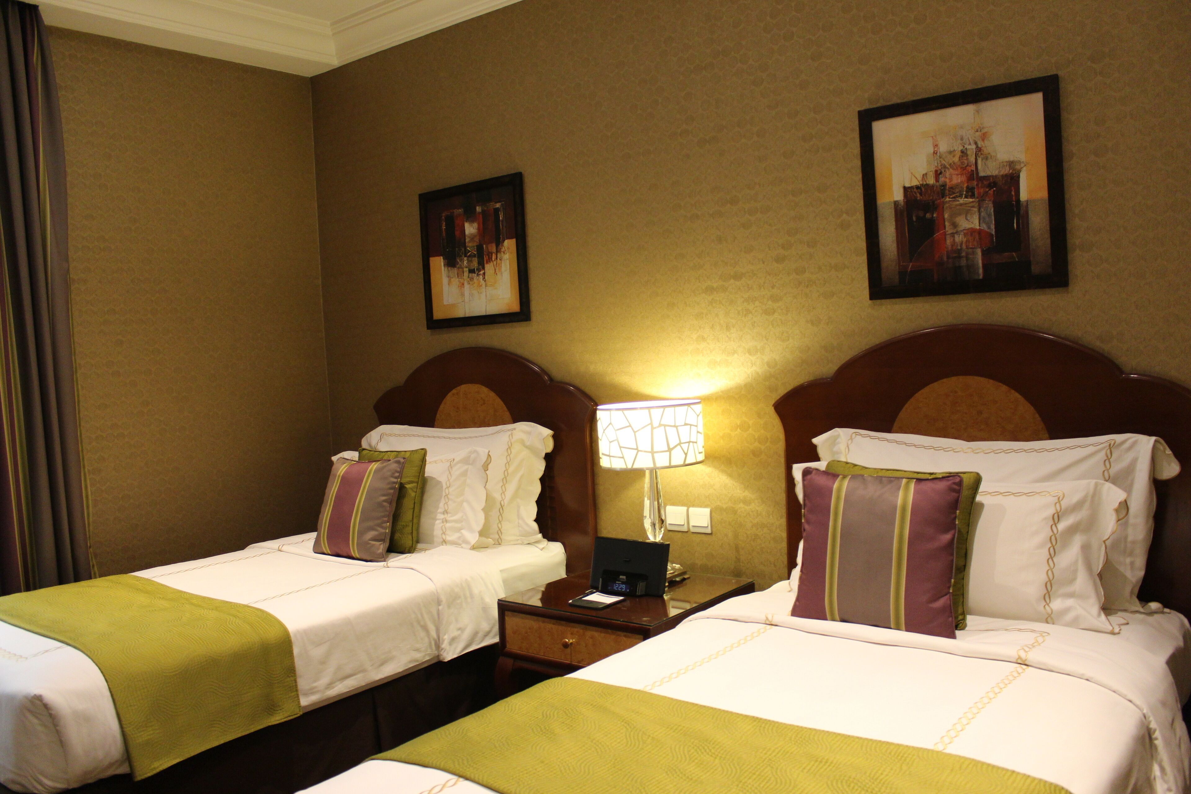 A hotel room with two single beds, each with a green bedspread, white linens, and decorative pillows. There is a nightstand with a lamp between the beds, and the walls are adorned with framed artwork.