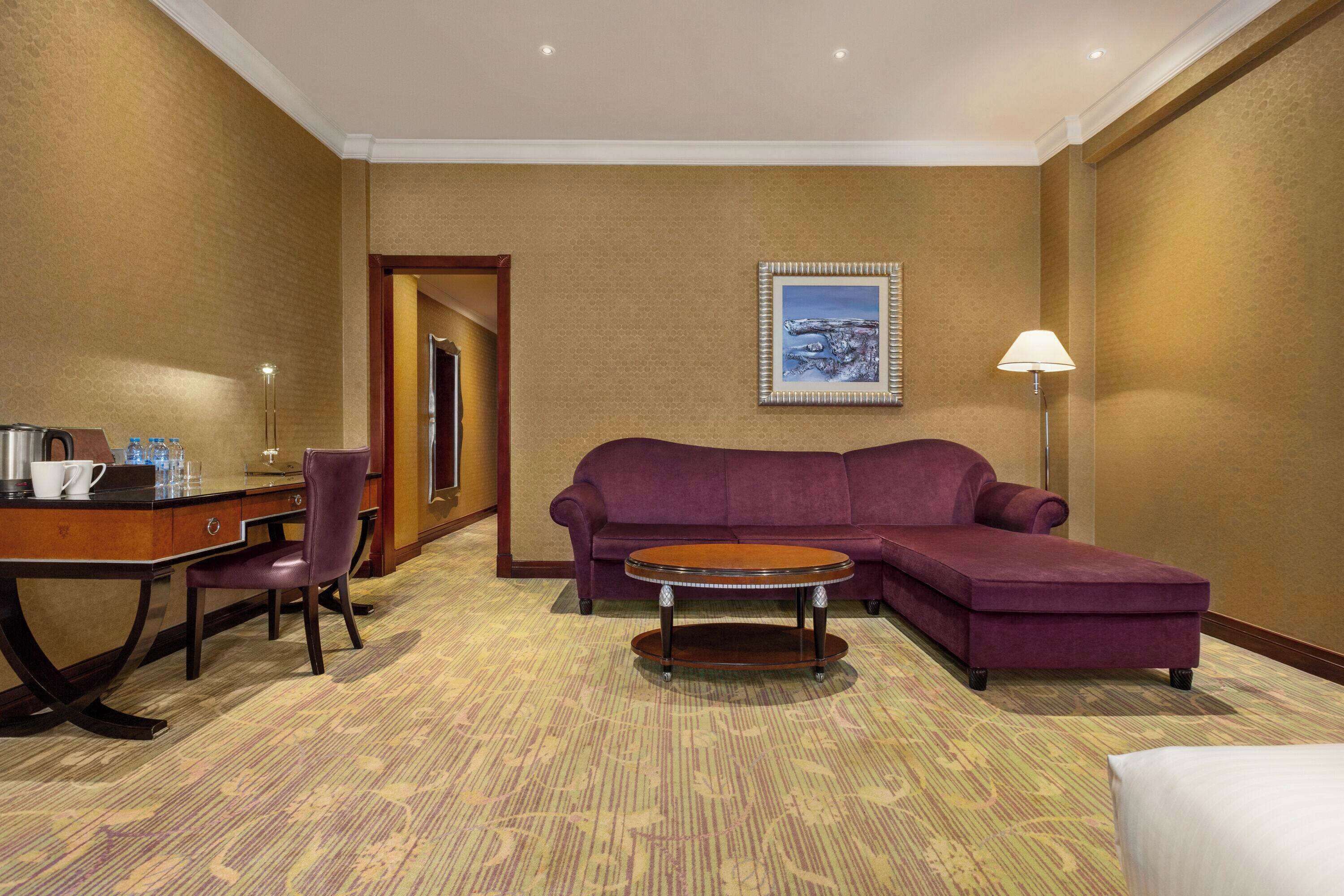 Quarto de hotel com sofá roxo, mesa de centro redonda, escrivaninha com cadeira, abajur e quadro na parede.