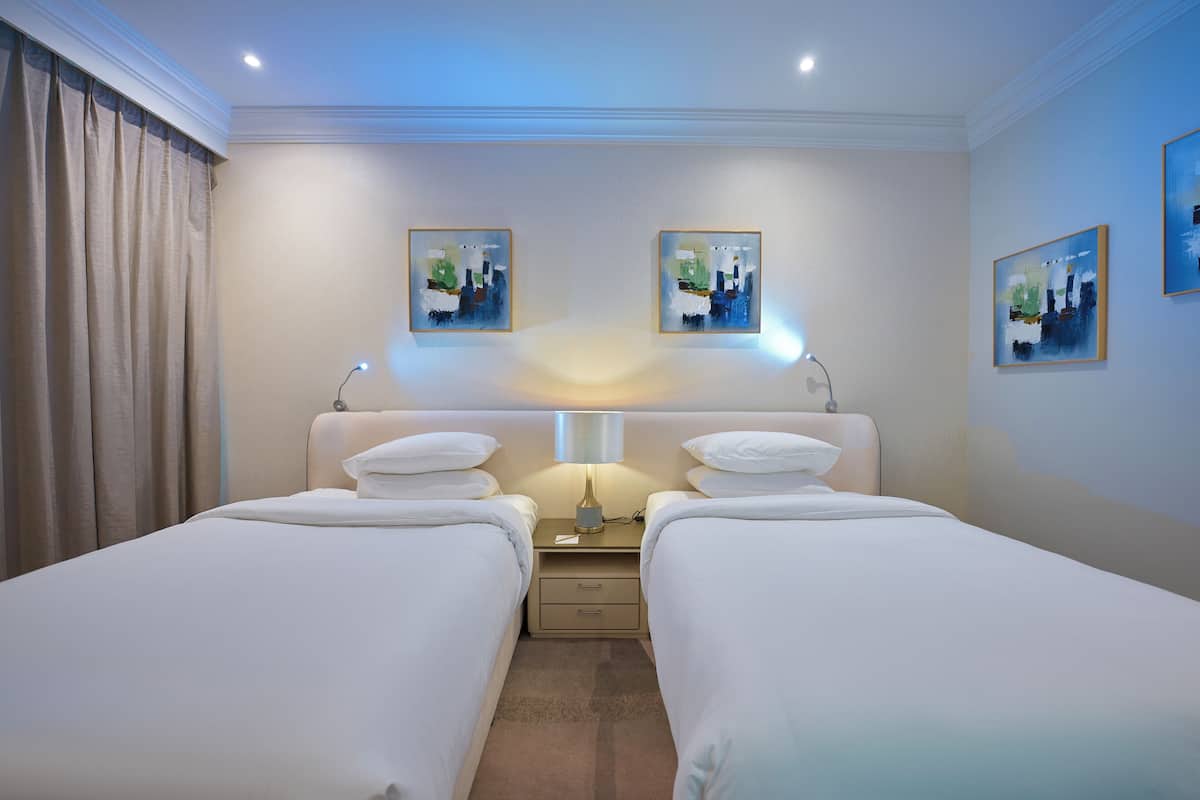 Habitación de hotel con dos camas, una mesita de noche, una lámpara y tres cuadros en la pared.