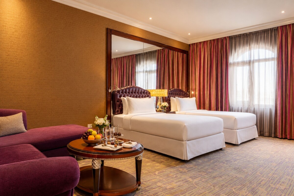A hotel room with a large bed, two bedside tables, a sofa, and a round coffee table with fruit and flowers.