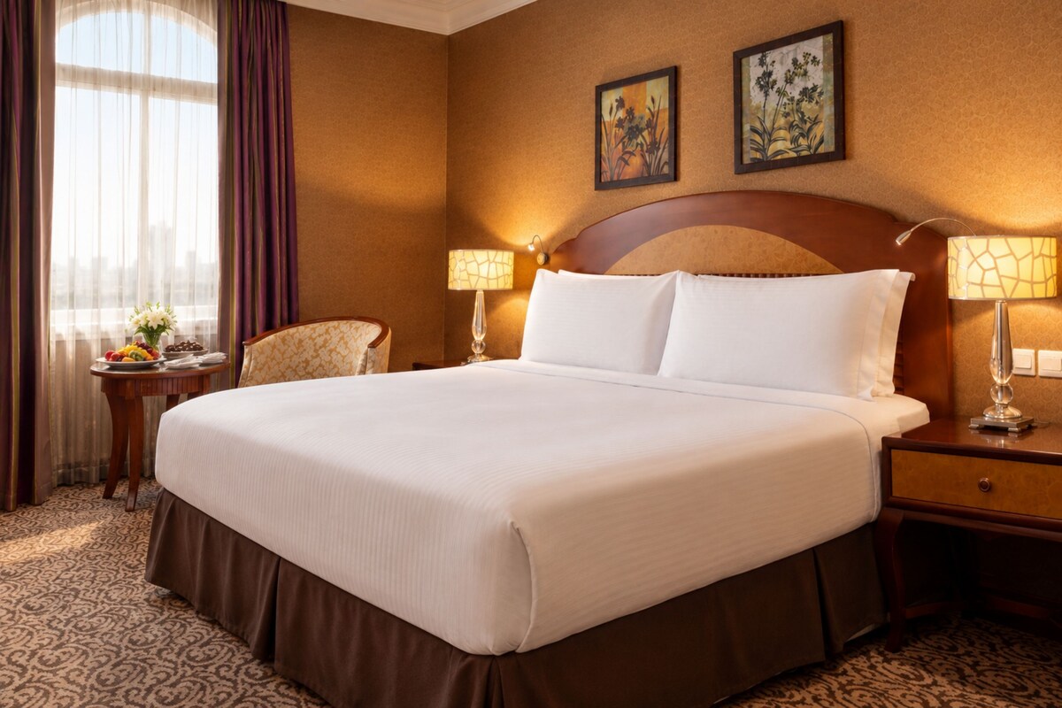 A hotel room with a large bed, two bedside lamps, a nightstand, a chair, a table with a vase of flowers, and two framed pictures on the wall.