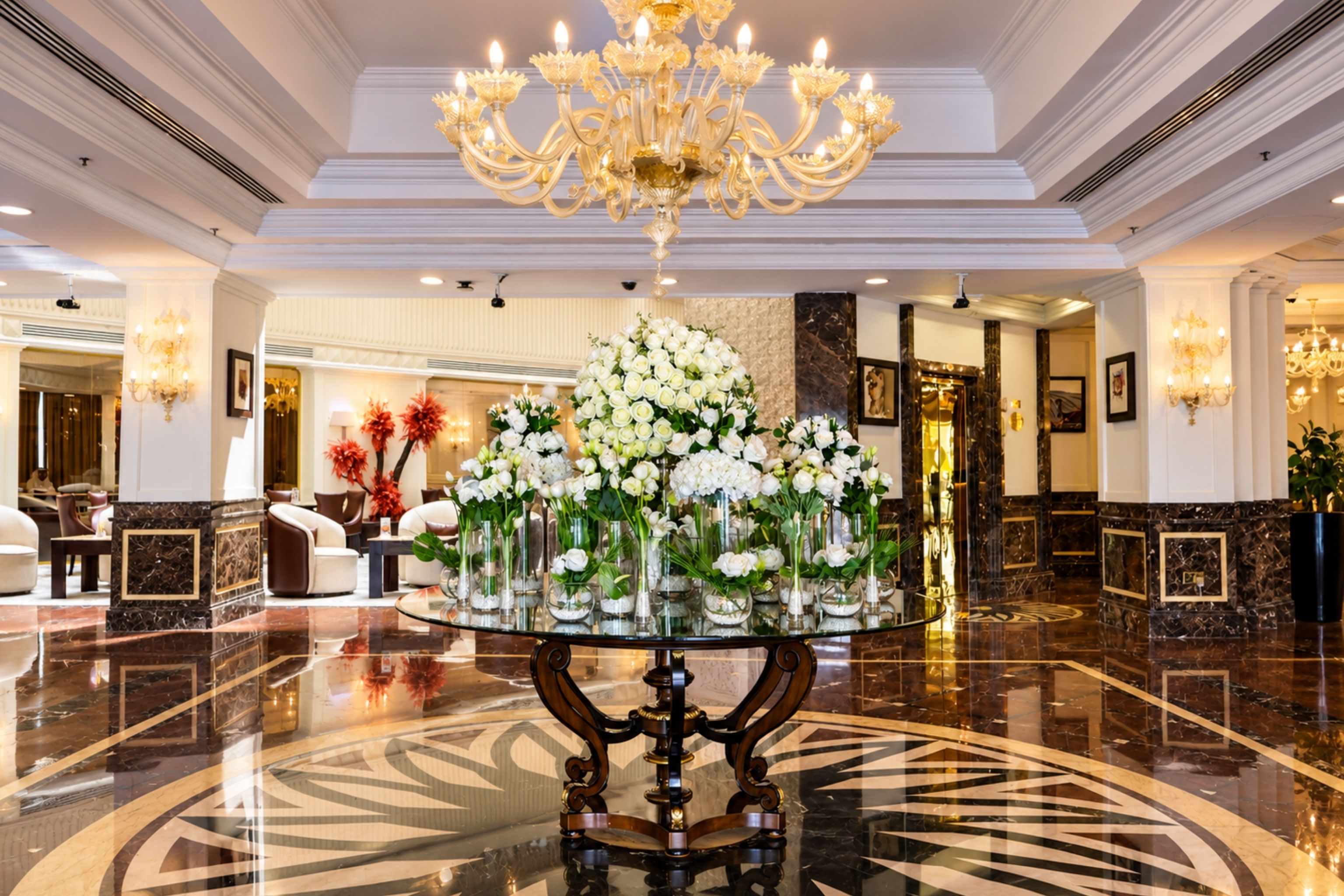 A lobby with a central table displaying floral arrangements, a chandelier, and marble flooring.