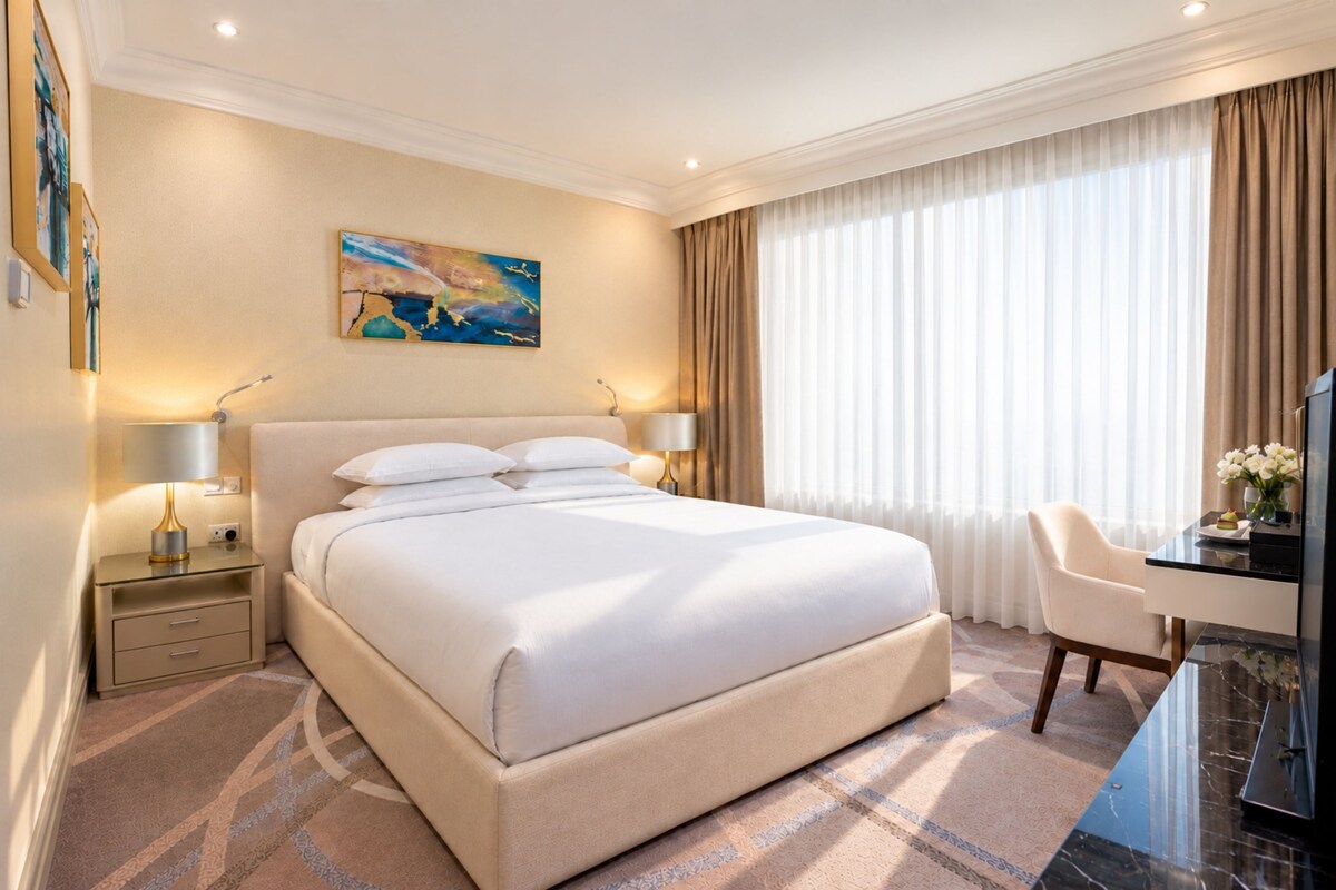 A hotel room with a large bed, bedside lamps, a desk with a chair, a painting on the wall, and a large window with curtains.