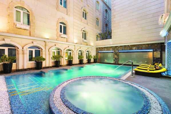 A swimming pool with a hot tub, surrounded by potted plants and a building with arched windows.