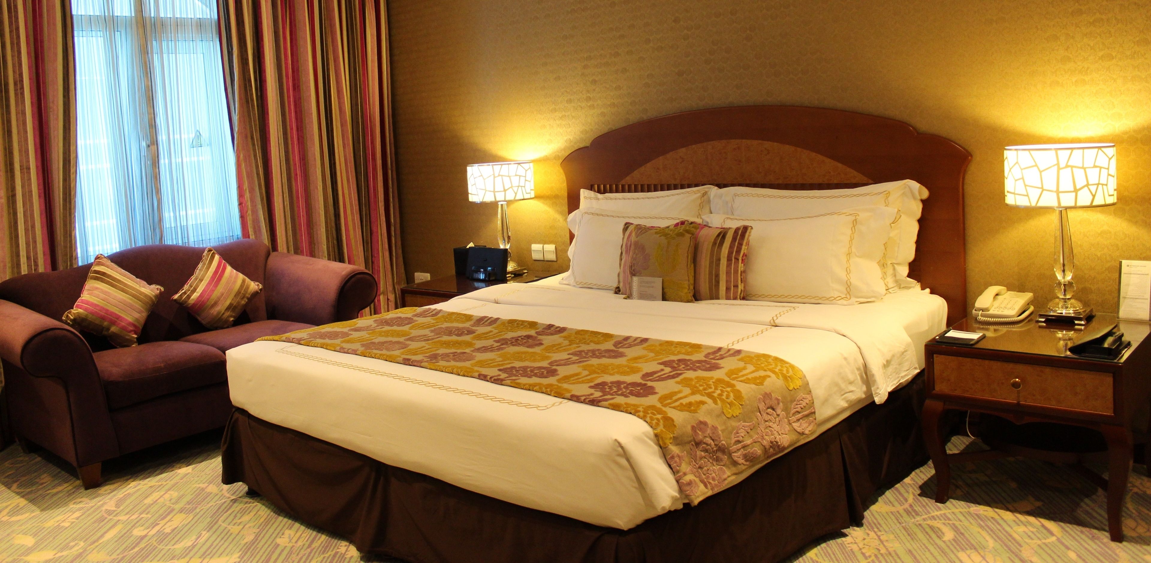 Quarto de hotel com uma cama grande, um sofá, duas luminárias de cabeceira e uma mesa de cabeceira com telefone e luminária.