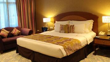 Quarto de hotel com uma cama grande, um sofá, duas luminárias de cabeceira e uma mesa de cabeceira com telefone e luminária.