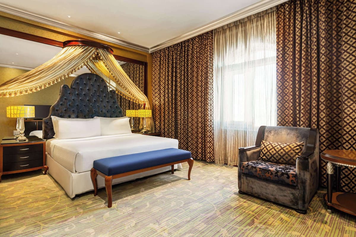 Una habitación de hotel con una cama grande, dosel, una silla, una mesita de noche y una lámpara.