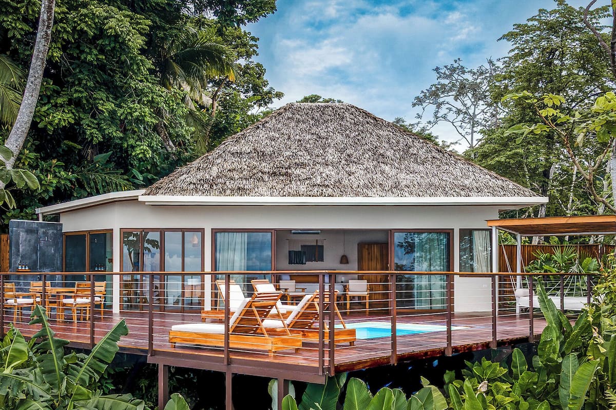 A modern house with a thatched roof, large windows, and a pool area with lounge chairs.