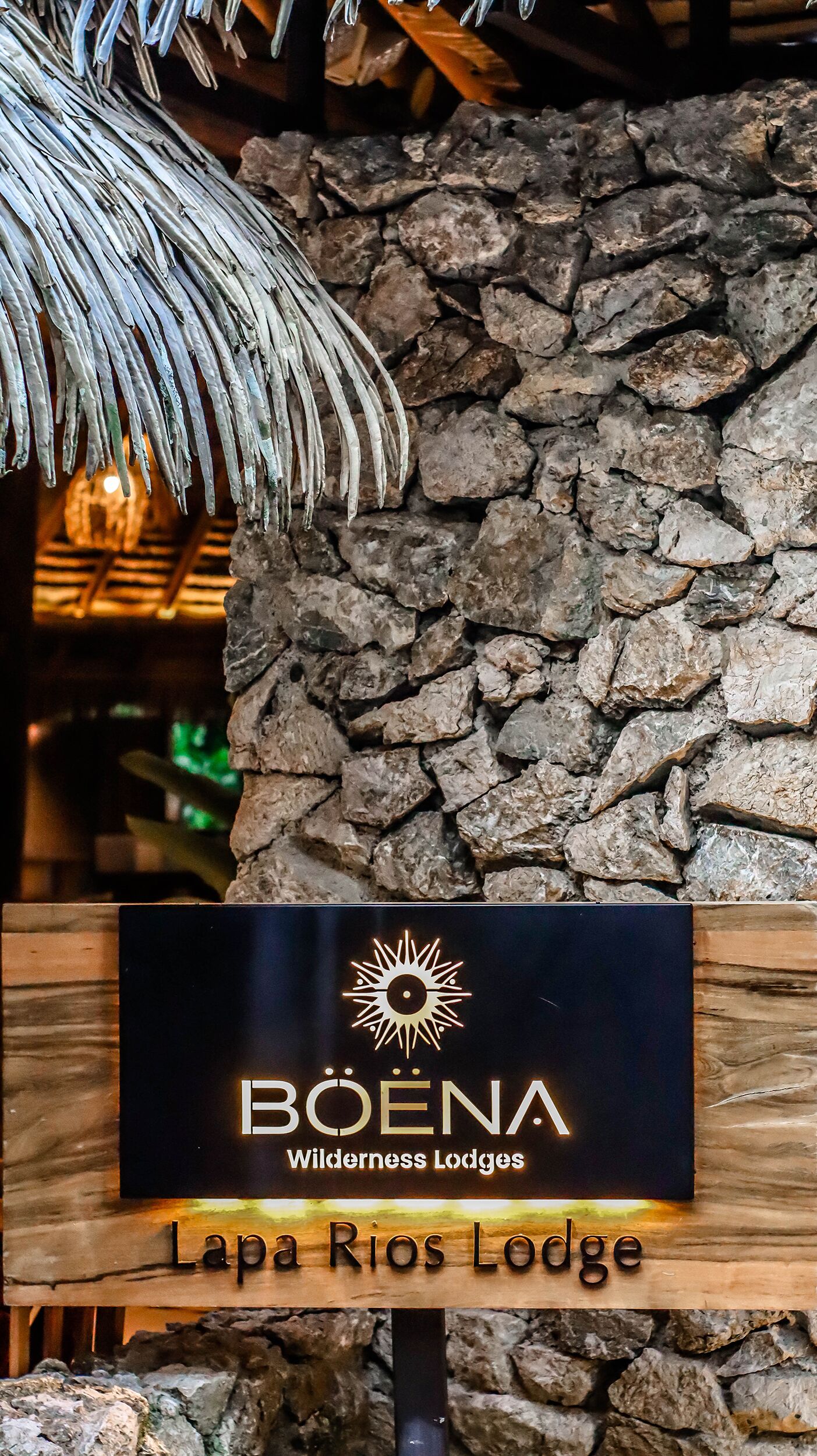 A sign for Boena Wilderness Lodges, Lapa Rios Lodge, with a rustic stone wall and a palm frond overhead.