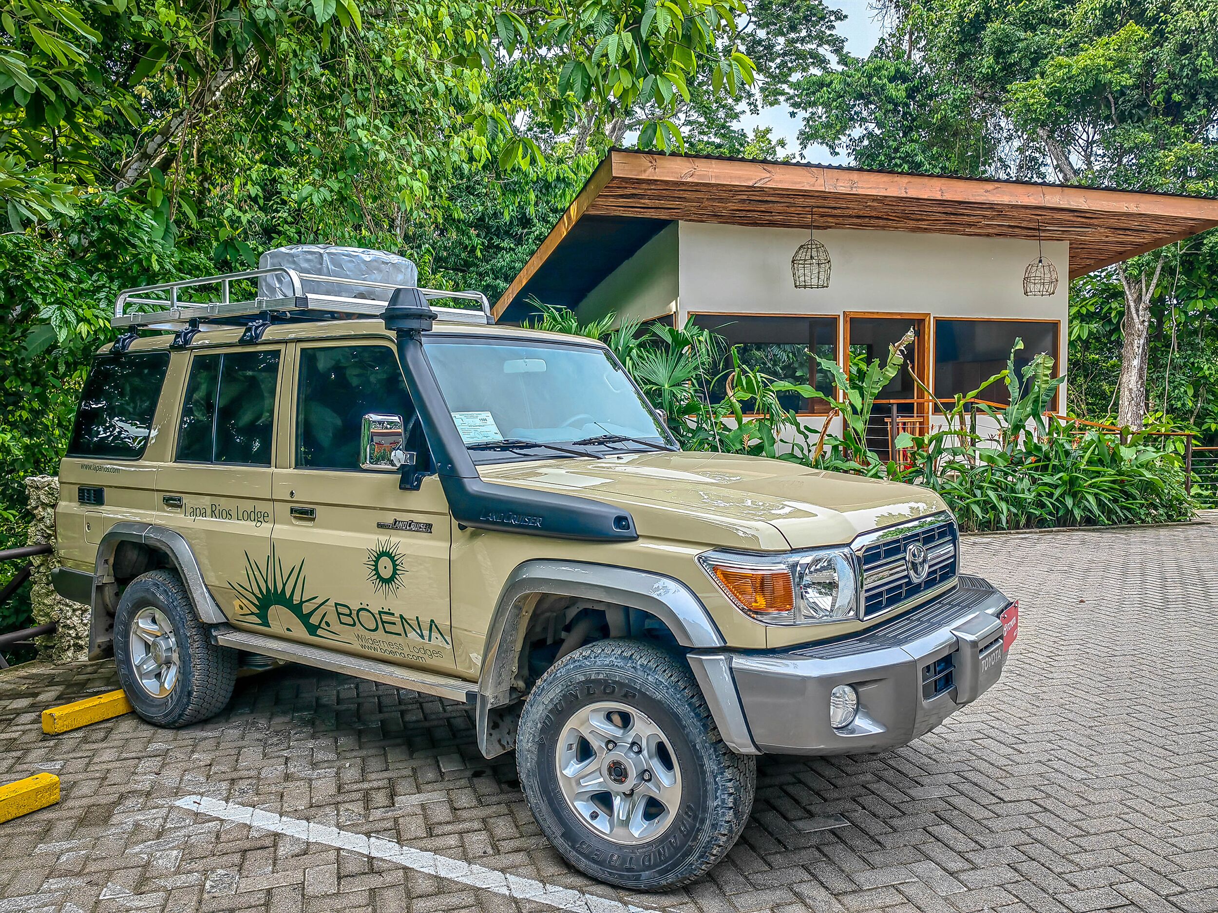A beige SUV parked in front of a building with a wooden roof and large windows, surrounded by lush greenery.