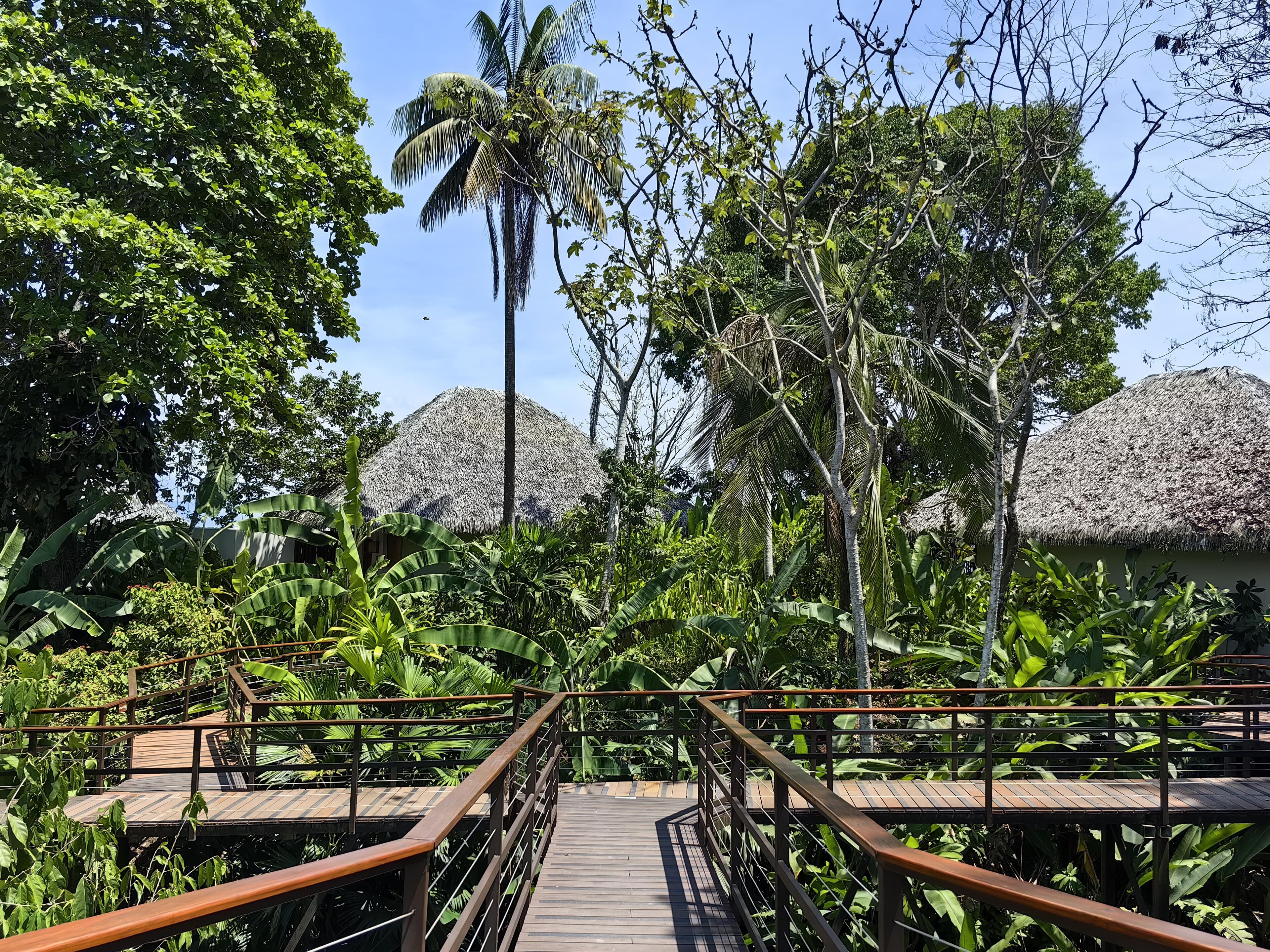 A wooden bridge leading to thatched-roof huts surrounded by lush tropical vegetation.