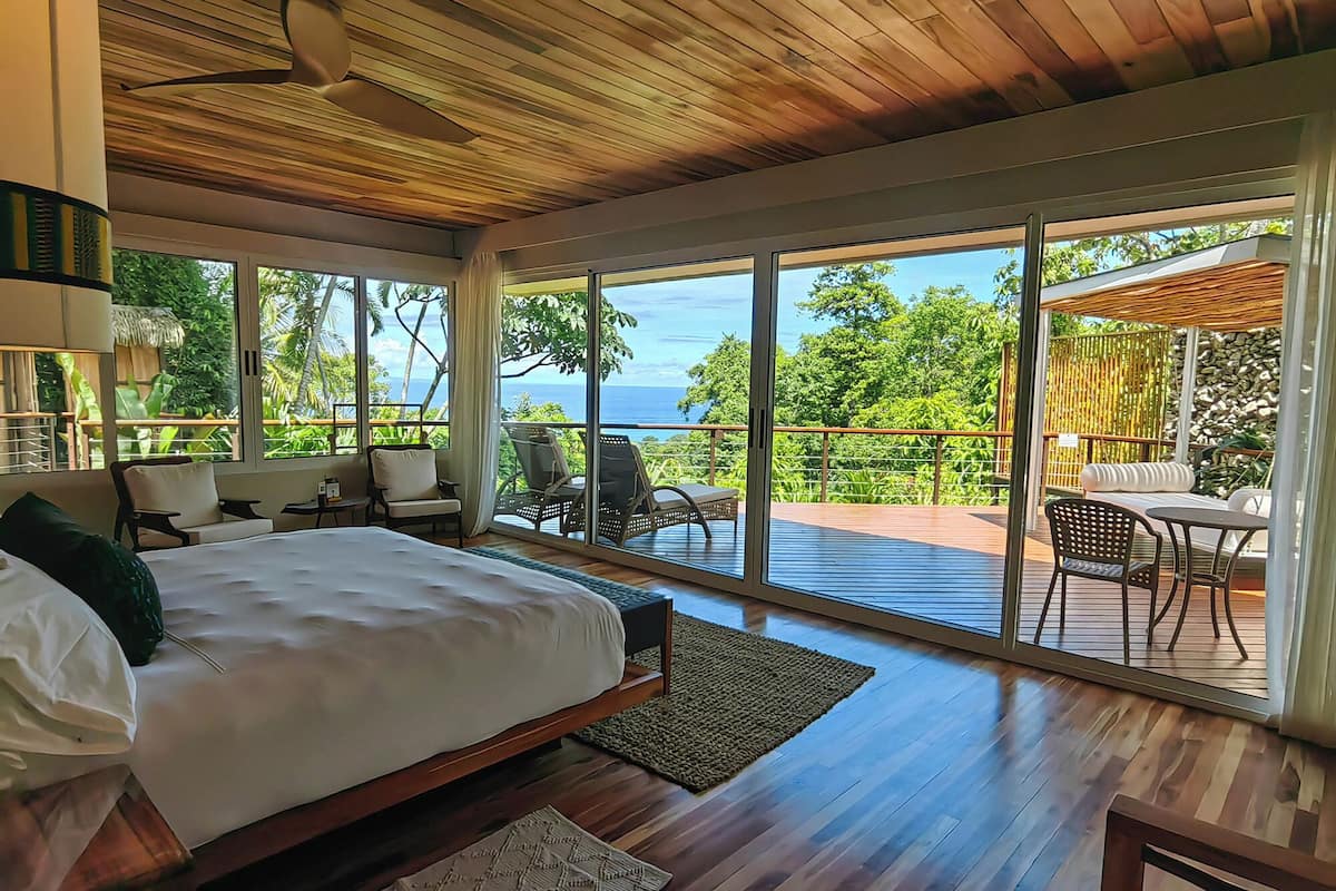 A spacious bedroom with a large bed, a wooden ceiling, and a balcony with a view of the ocean.