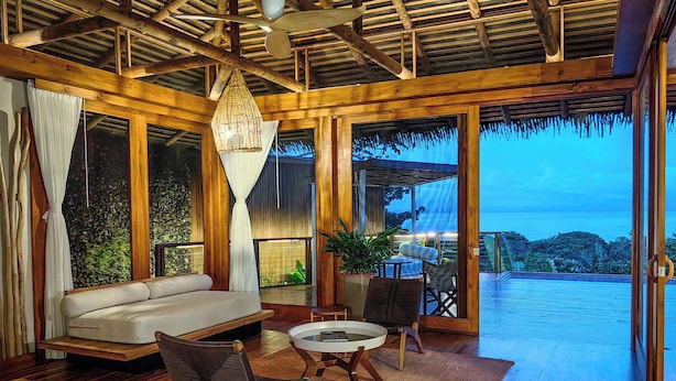A spacious room with a wooden ceiling, large windows, a sofa, chairs, and a small table. - Lapa Rios Lodge (Puerto Jiménez)