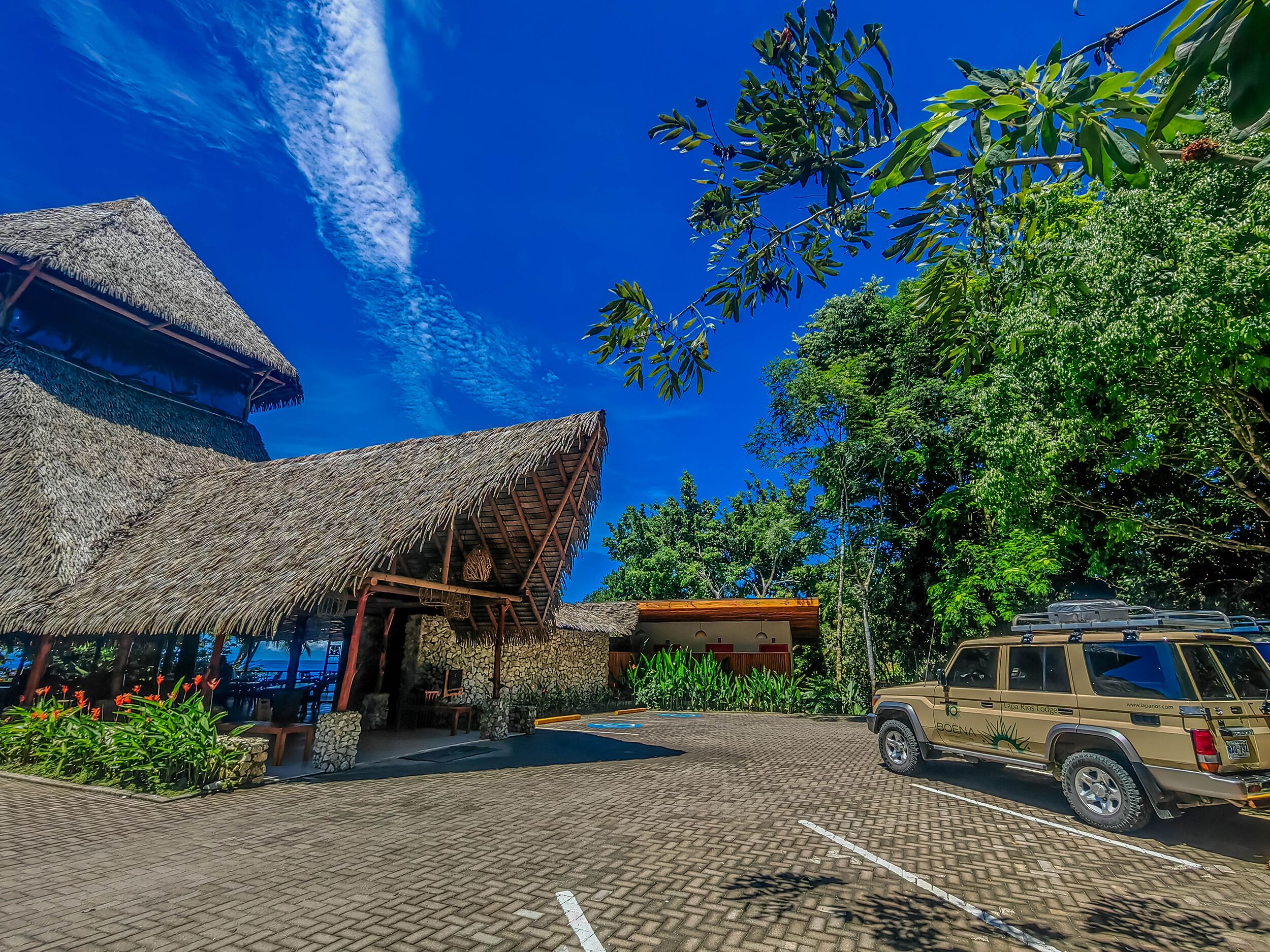 A thatched-roof building with a paved parking area and a parked SUV.