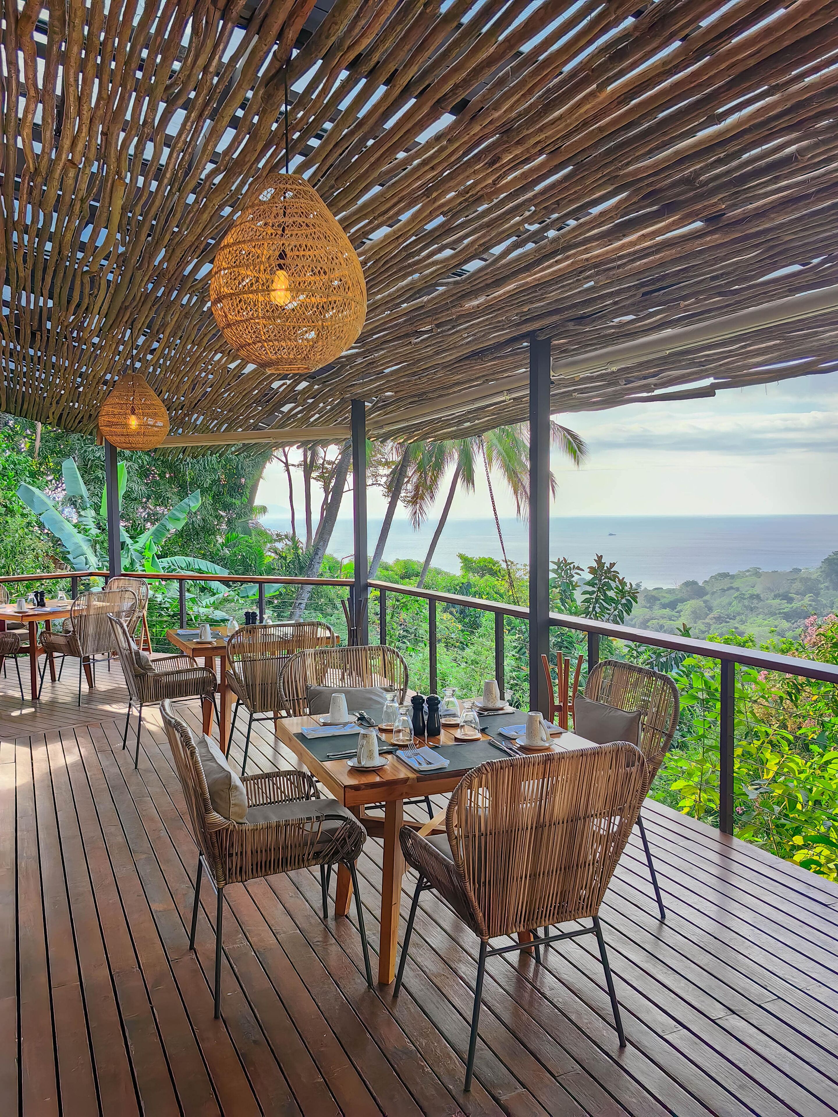 A wooden deck with wicker furniture and hanging lights, overlooking a lush landscape and the sea.