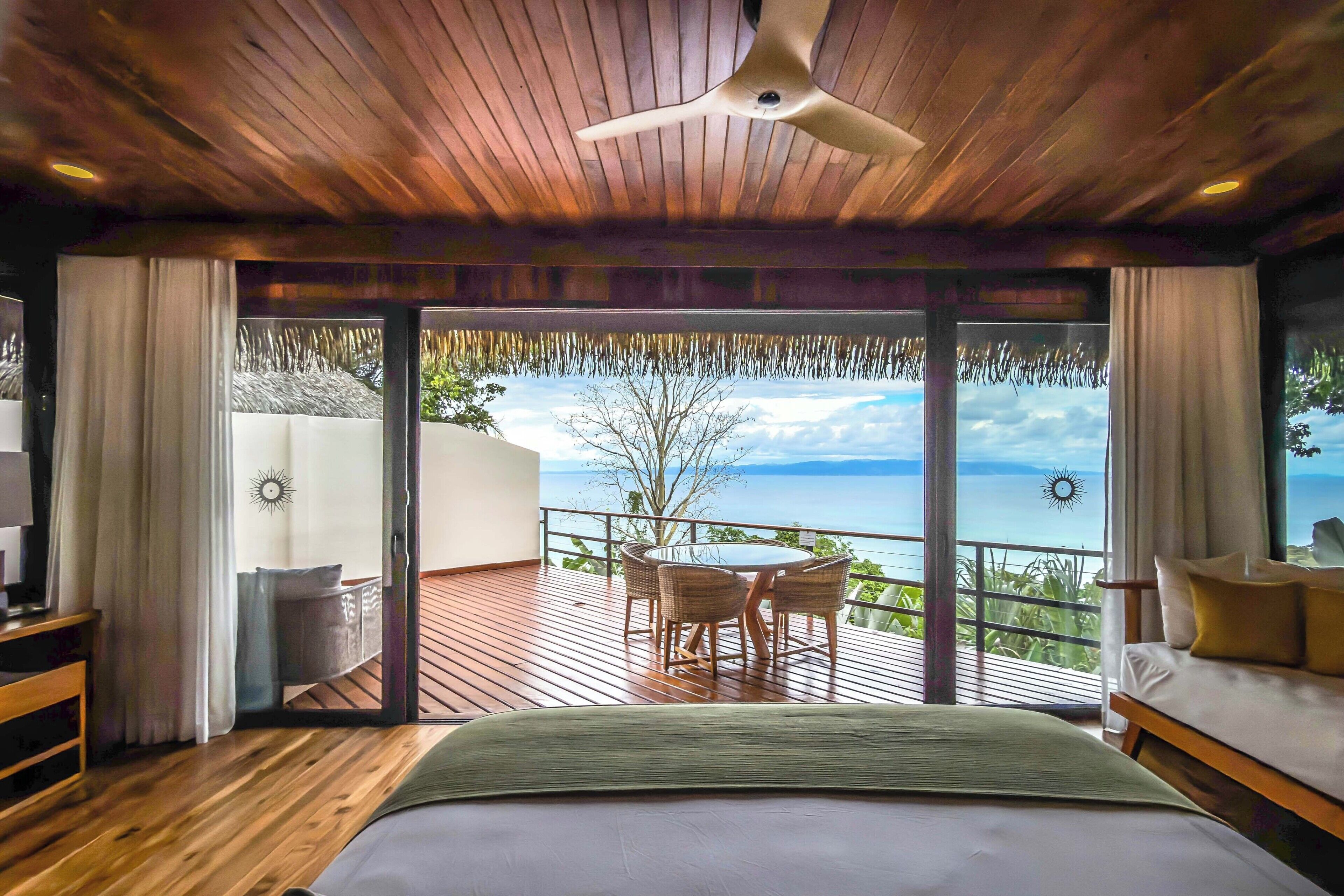 Un dormitorio con techo de madera, una cama, un balcón con mesa y sillas, y vista al mar.