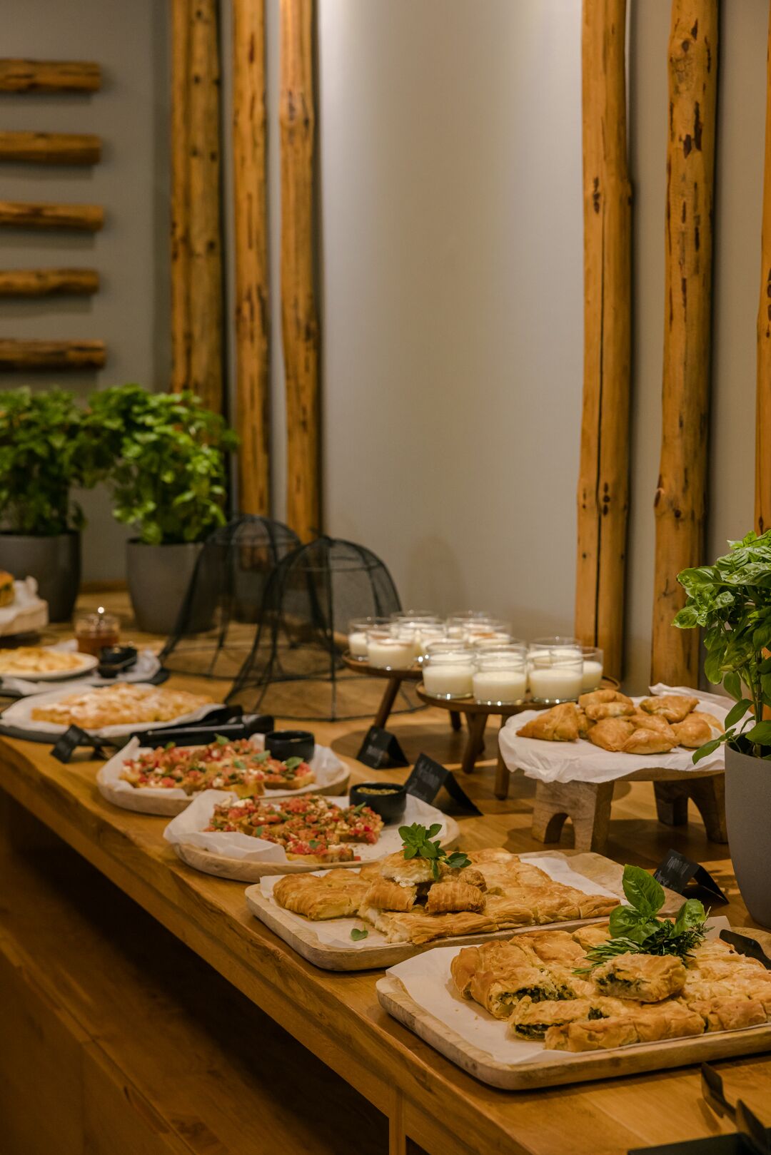 Une table de buffet proposant divers plats, notamment des pâtisseries et des salades, dans une pièce agrémentée de boiseries et de plantes en pot.