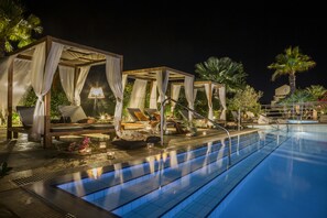 A poolside area with a clear blue pool, wooden cabanas with white drapes, lounge chairs, and ambient lighting. - Avithos Resort Apartments Hotel (Kefalonia)