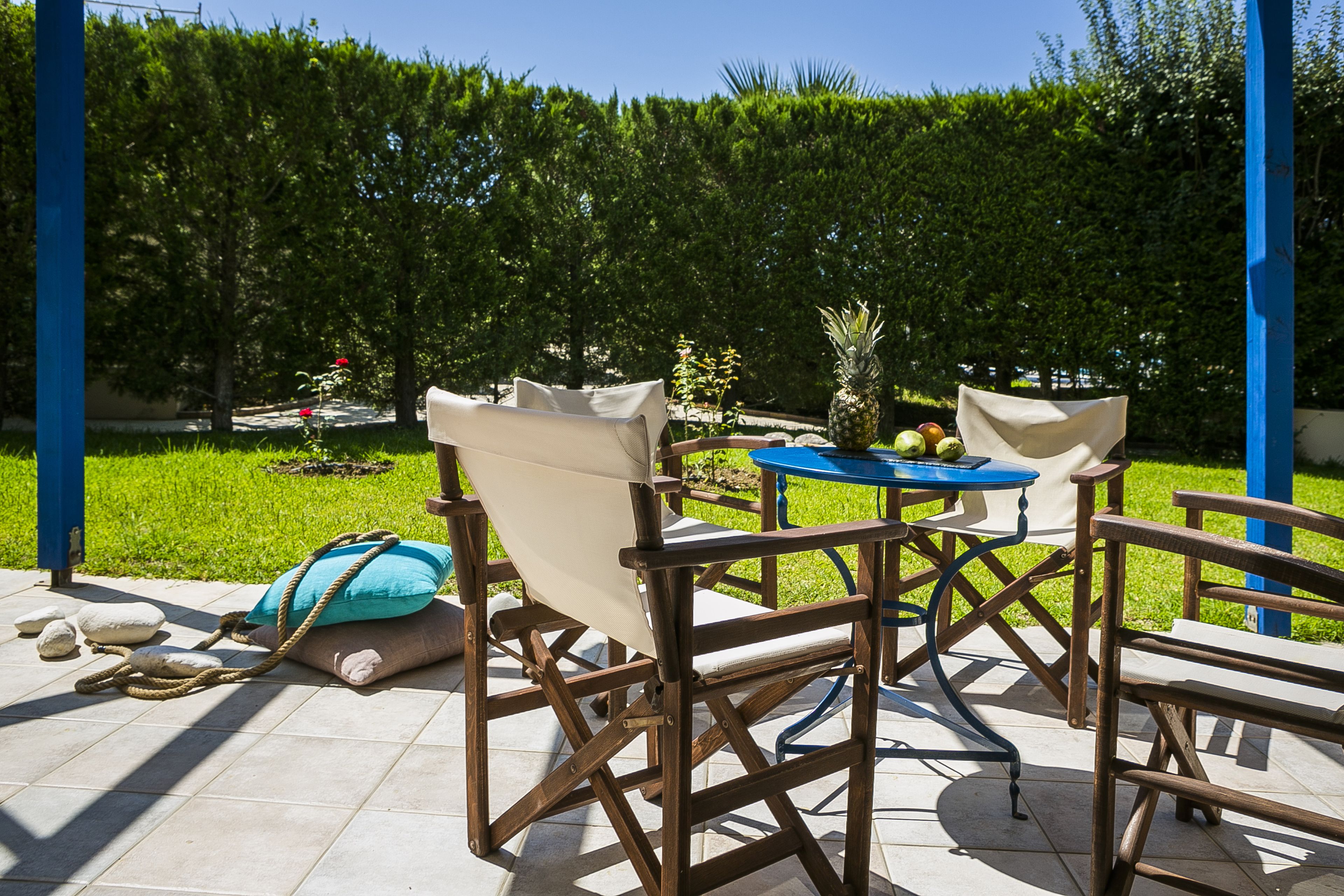 An outdoor seating area with a table and chairs, surrounded by a hedge and a blue pillar.