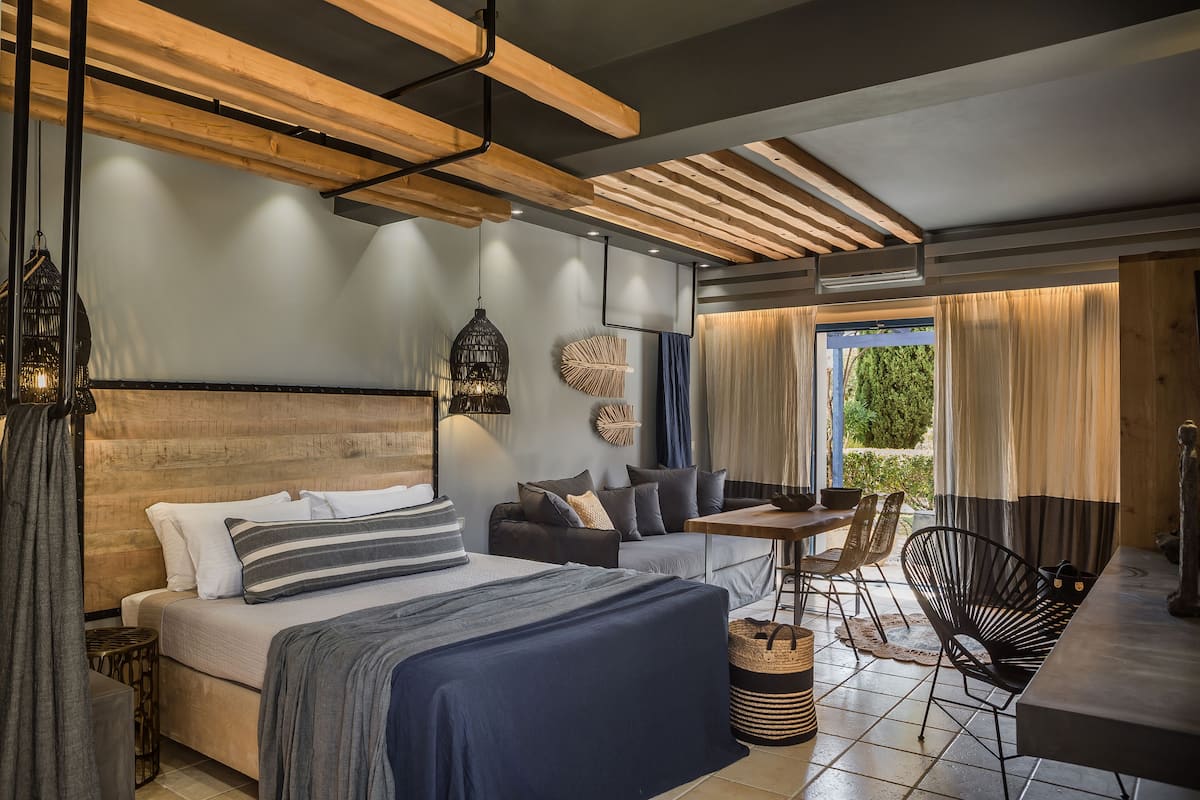 A modern hotel room with a large bed, a sofa, a dining table, and a chair. There is a window with curtains and a view of greenery outside.
