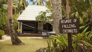 Un panneau d’avertissement indiquant un risque de chute de noix de coco, des palmiers et une maison avec une véranda couverte.