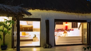 Un hôtel dont l’entrée est ouverte et mène à un intérieur bien éclairé, avec un toit de chaume et une terrasse en bois.
