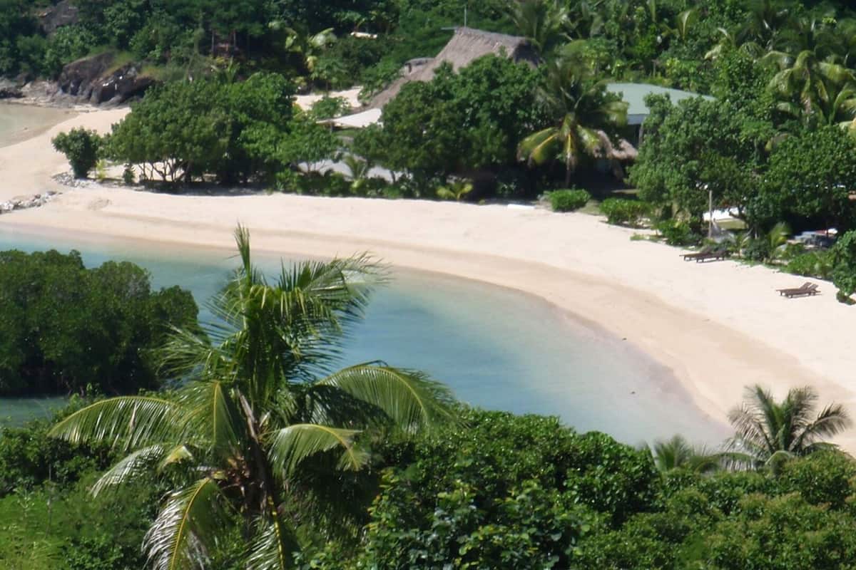 A beach with clear turquoise water, surrounded by lush greenery and a few buildings.