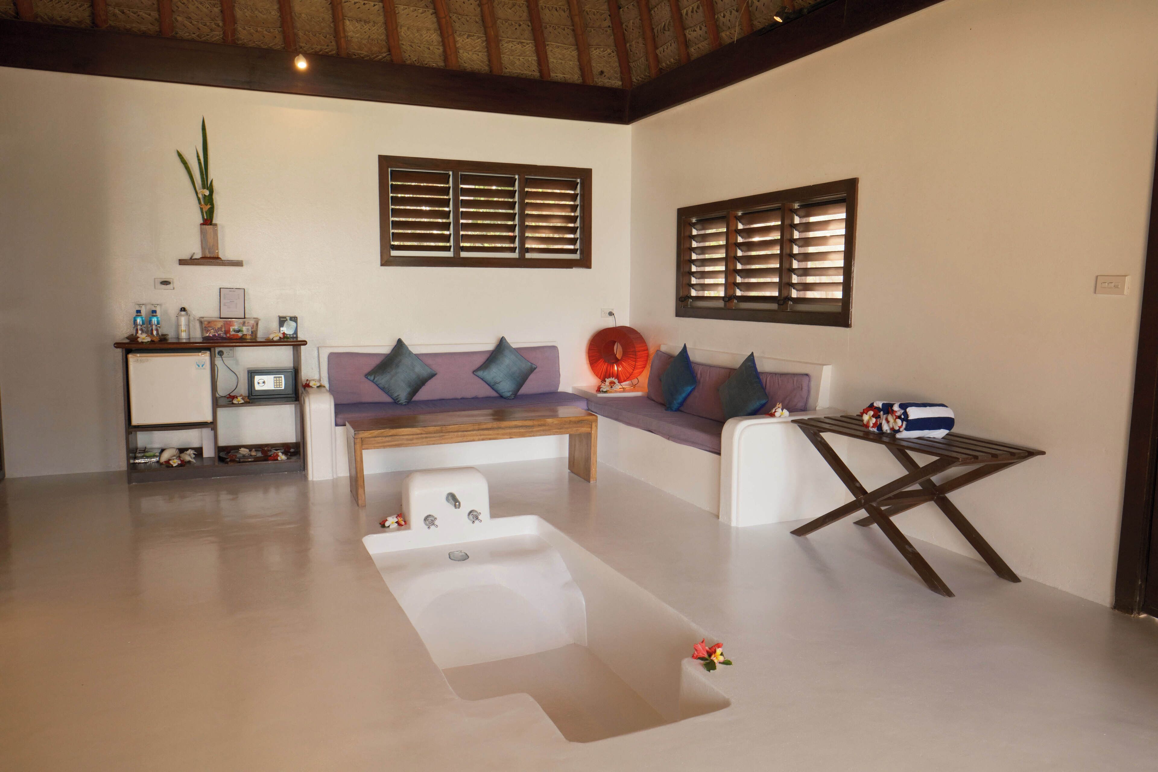 A modern living room with a sofa, a coffee table, and a small table with a bag. There is a plant on a shelf and a window with blinds.