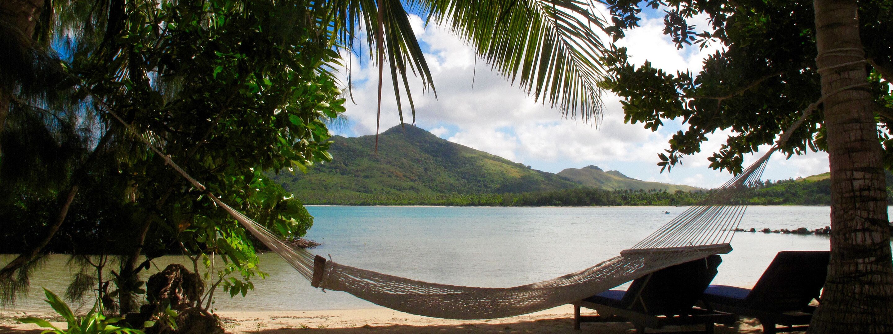 A hammock tied between trees on a beach with a view of a mountain and calm water.