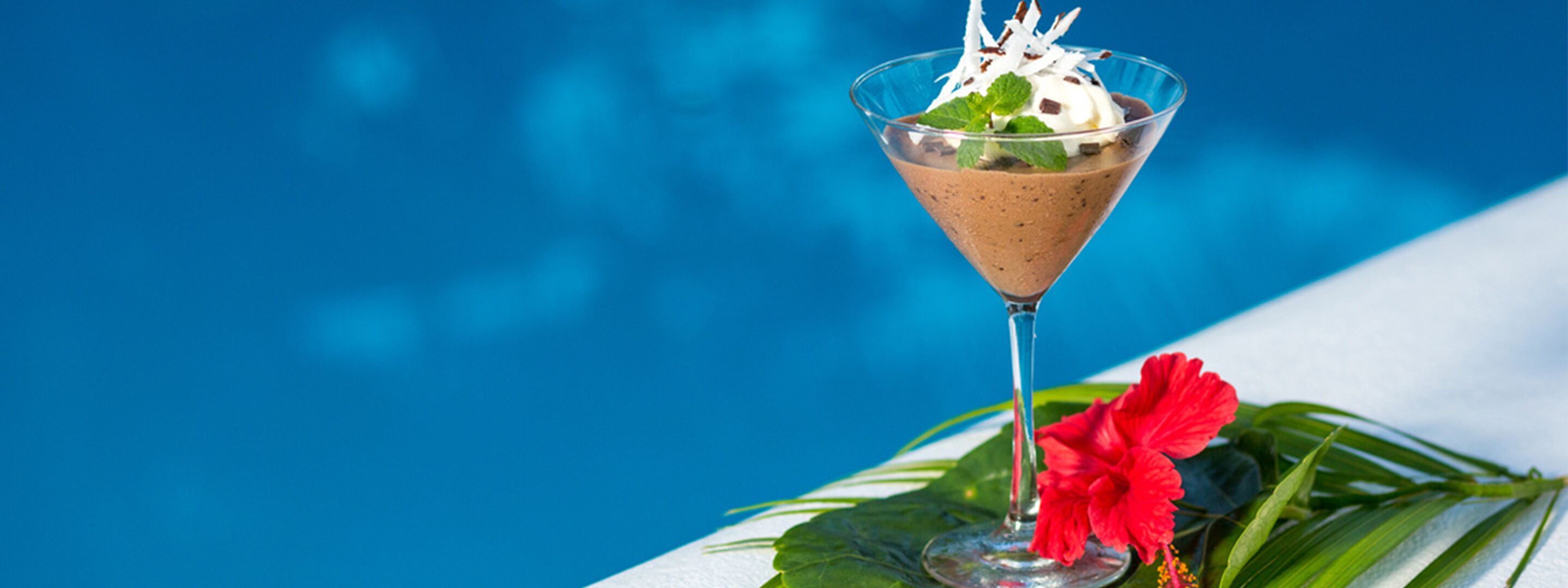 A chocolate dessert in a martini glass with whipped cream and mint leaves, placed on a table with a red hibiscus flower and green leaves, against a blue background.