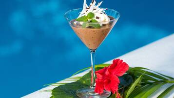 A chocolate dessert in a martini glass with whipped cream and mint leaves, placed on a table with a red hibiscus flower and green leaves, against a blue background.