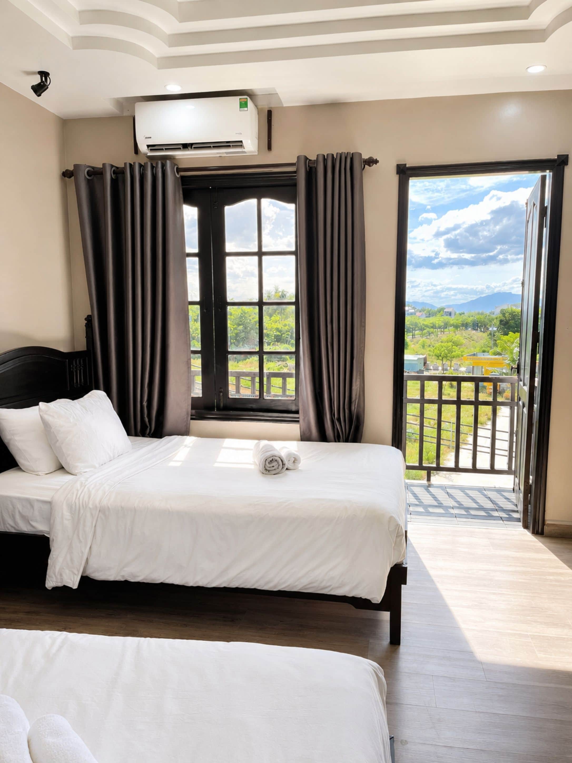 A hotel room with two beds, a large window with curtains, an air conditioning unit, and a view of greenery and buildings outside.