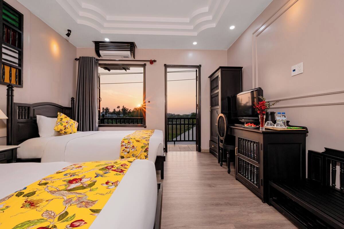 A hotel room with two beds, a TV, a minibar, and a balcony with a sunset view.
