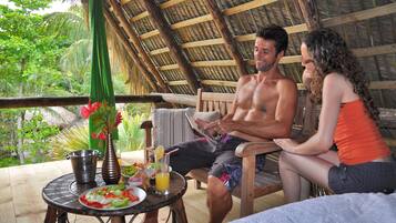 A couple sitting on a wooden bench under a thatched roof, with a table in front of them holding a plate of food, a drink, and a vase with flowers.